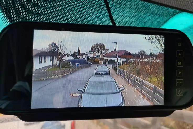 Display eines digitalen R&uuml;ckspiegels zeigt Stra&szlig;enbild mit zwei Autos, H&auml;usern, Gehweg, Zaun und B&auml;umen unter bew&ouml;lktem Himmel