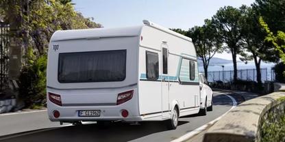 Hinteransicht eines wei&szlig;en Wohnwagens, an einen Pkw angeschlossen, auf einer K&uuml;stenstra&szlig;e; rechts Steinbr&uuml;stung, links B&auml;ume, Wasser im Hintergrund