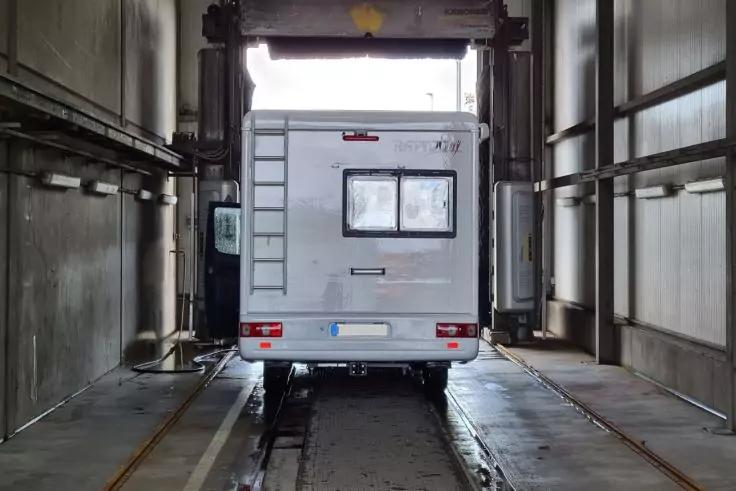 R&uuml;ckansicht eines wei&szlig;en Wohnmobils auf Schienen in einem schmalen, hohen Raum mit Metallw&auml;nden und heller &Ouml;ffnung hinten