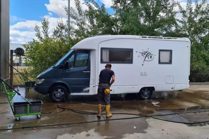 Person w&auml;scht wei&szlig;es Wohnmobil im Au&szlig;enbereich mit Schlauch; Pf&uuml;tzen, B&auml;ume und blauer Himmel mit Wolken im Hintergrund