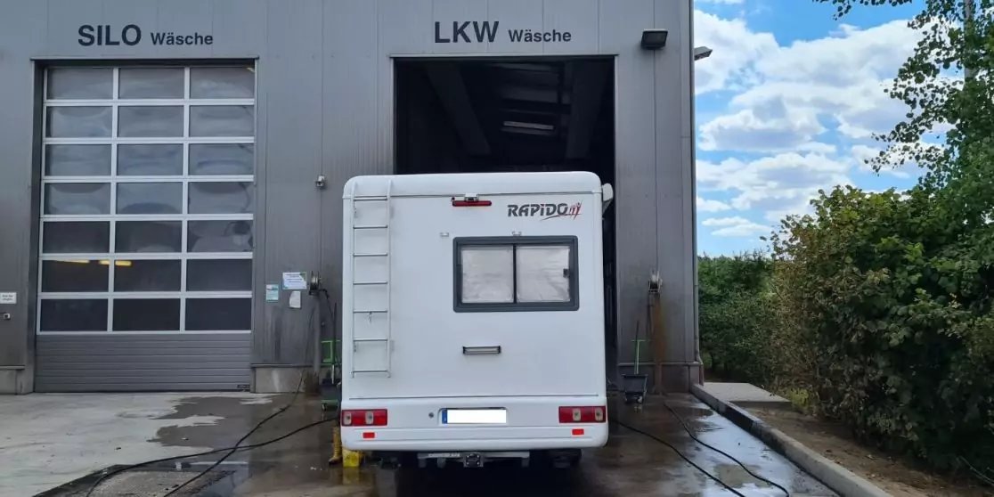 R&uuml;ckansicht eines wei&szlig;en Wohnmobils vor zwei Waschboxen mit den Schildern 'SILO W&auml;sche' und 'LKW W&auml;sche', nasser Boden, Heckfenster, B&auml;ume rechts, bew&ouml;lkter Himmel