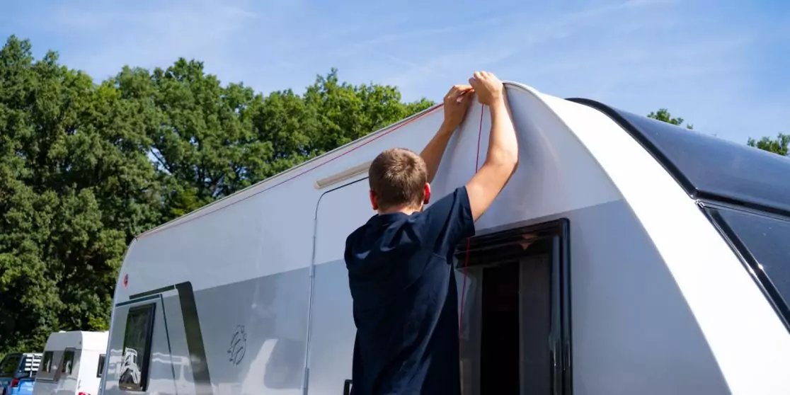 Person h&auml;lt aufgerollte Markise am Dach eines Wohnwagens; B&auml;ume und blauer Himmel im Hintergrund