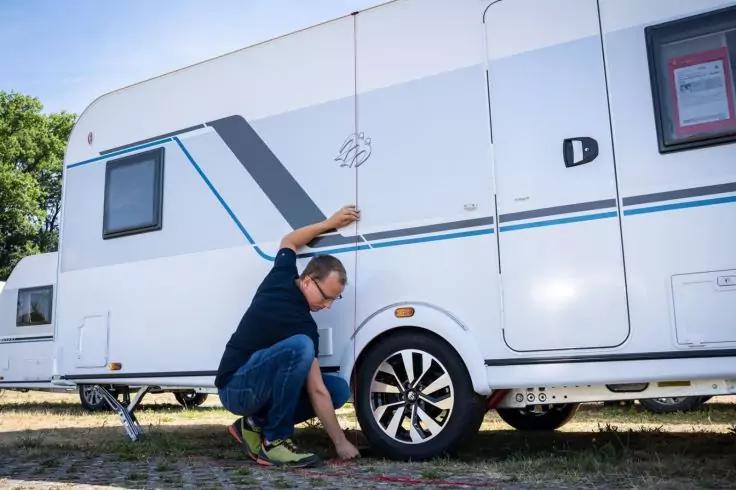 Eine Person hockt neben dem rechten Rad eines wei&szlig;en Wohnwagens und ber&uuml;hrt den Reifen