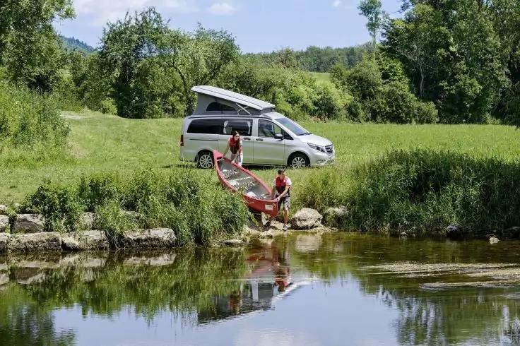 Wei&szlig;er Campingbus mit aufgeklapptem Dach auf einer grasigen Flussuferwiese; zwei Personen tragen ein rotes Kanu ins Wasser; B&auml;ume im Hintergrund