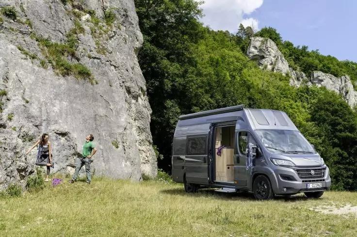 Kastenwagen mit ge&ouml;ffneter Seitent&uuml;r auf einer Wiese neben einer Felswand, zwei Personen nahe den Felsen, B&auml;ume und Himmel im Hintergrund