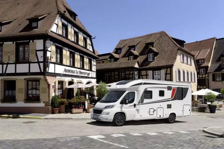 Wei&szlig;es Reisemobil auf gepflastertem Platz vor Fachwerkh&auml;usern mit Au&szlig;engastronomie und blauem Himmel