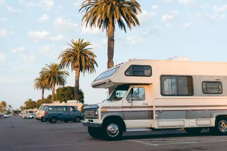 Wei&szlig;es Wohnmobil auf einem Parkplatz, Palmen im Hintergrund, blauer Himmel mit Wolken, weitere geparkte Fahrzeuge