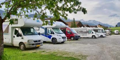 Reihe geparkter Wohnmobile auf einem Kiesplatz, Baumzweig links, H&auml;user und Berge im Hintergrund, bew&ouml;lkter Himmel