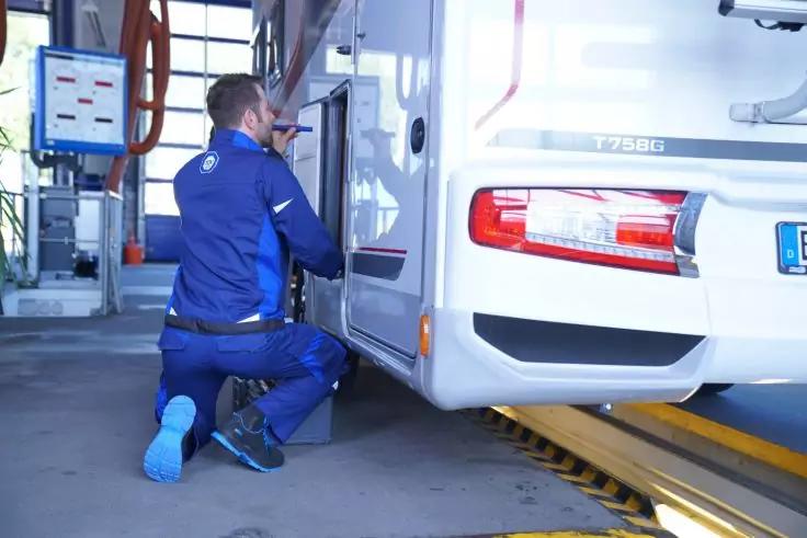 Person in blauem Arbeitsanzug kniet neben einem wei&szlig;en Wohnmobil in einer Werkstatt und &ouml;ffnet eine Seitenklappe; Hebeb&uuml;hne und Monitor im Hintergrund