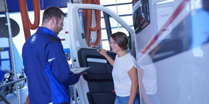 Mann in blauem Arbeitsanzug h&auml;lt Tablet neben ge&ouml;ffneter Seitent&uuml;r eines wei&szlig;en Fahrzeugs; Frau in wei&szlig;em T‑Shirt steht im Einstieg; orangefarbene Schl&auml;uche und Werkstattausr&uuml;stung im Hintergrund