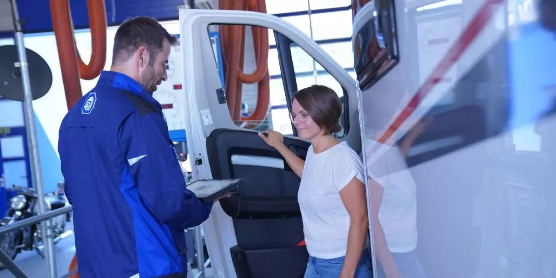 Mann in blauem Arbeitsanzug h&auml;lt Tablet neben ge&ouml;ffneter Seitent&uuml;r eines wei&szlig;en Fahrzeugs; Frau in wei&szlig;em T‑Shirt steht im Einstieg; orangefarbene Schl&auml;uche und Werkstattausr&uuml;stung im Hintergrund