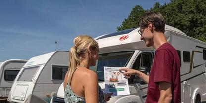 Ein Mann und eine Frau stehen vor einem Wohnwagen; der Mann zeigt auf ein an der Fahrzeugfront befestigtes Prospekt. Im Hintergrund weitere Wohnwagen, B&auml;ume und blauer Himmel