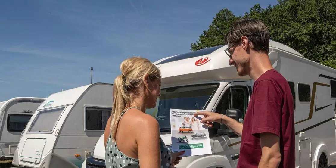 Ein Mann und eine Frau stehen vor einem Wohnwagen; der Mann zeigt auf ein an der Fahrzeugfront befestigtes Prospekt. Im Hintergrund weitere Wohnwagen, B&auml;ume und blauer Himmel