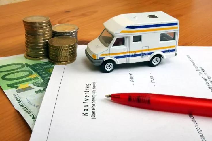 Modell-Wohnmobil auf einem Dokument mit der &Uuml;berschrift "Kaufvertrag", roter Stift darauf, gestapelte Eurom&uuml;nzen und teilweise sichtbarer 100-Euro-Schein auf einer Holzfl&auml;che