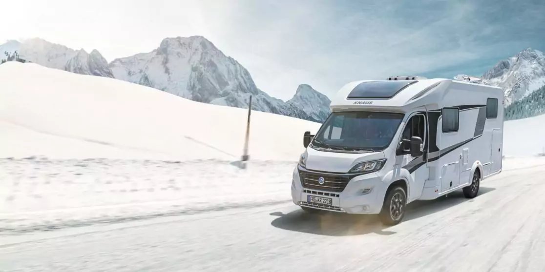 Wei&szlig;es Wohnmobil auf schneebedeckter Stra&szlig;e, im Hintergrund schneebedeckte Berge und blauer Himmel mit Wolken