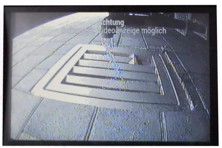 Bild eines Bildschirms mit einer Kameraufnahme eines quadratischen, gestuften Metallgitters im Pflaster; Pflastersteine darum; lineare Markierungen rechts; oben wei&szlig;e Einblendung &sbquo;Achtung Videoanzeige m&ouml;glich&lsquo;