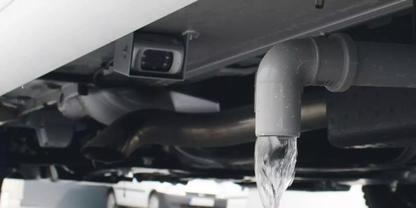 Graues, gebogenes Rohr an der Unterseite eines Fahrzeugs oder einer Maschine, klares Wasser flie&szlig;t als schmaler Strahl heraus; im Hintergrund dunkle Metallrohre