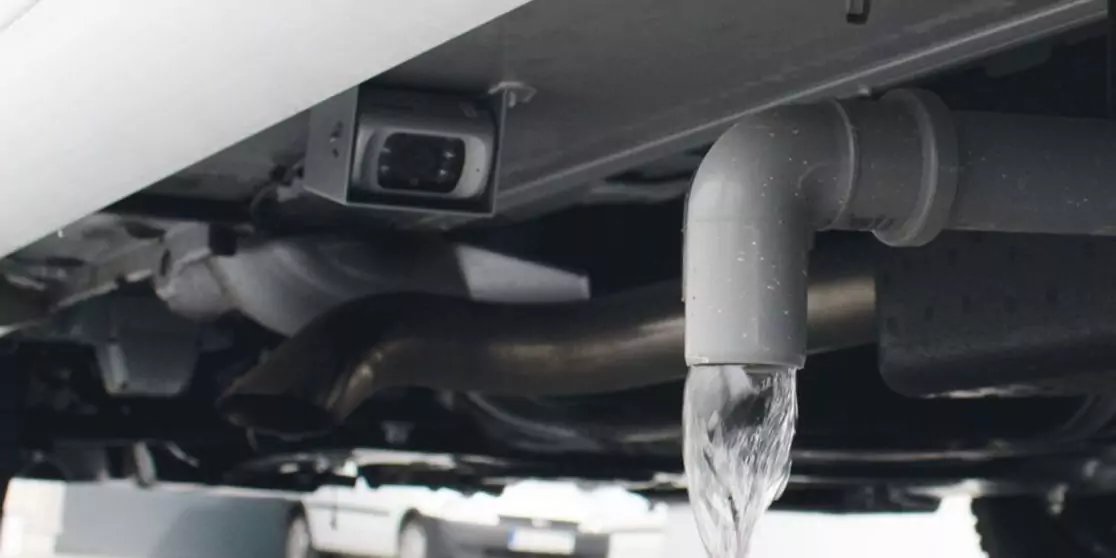 Graues, gebogenes Rohr an der Unterseite eines Fahrzeugs oder einer Maschine, klares Wasser flie&szlig;t als schmaler Strahl heraus; im Hintergrund dunkle Metallrohre