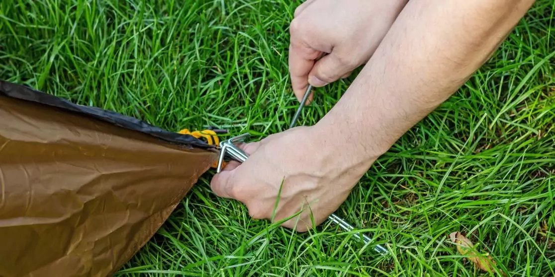 Zwei H&auml;nde befestigen ein Metallteil an der Kante eines braunen Zeltes auf gr&uuml;nem Gras