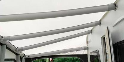 Blick unter wei&szlig;es Markisendach mit gebogenen Metallstreben, Seitenwand mit Fenster und T&uuml;r, gr&uuml;ne B&auml;ume im Hintergrund