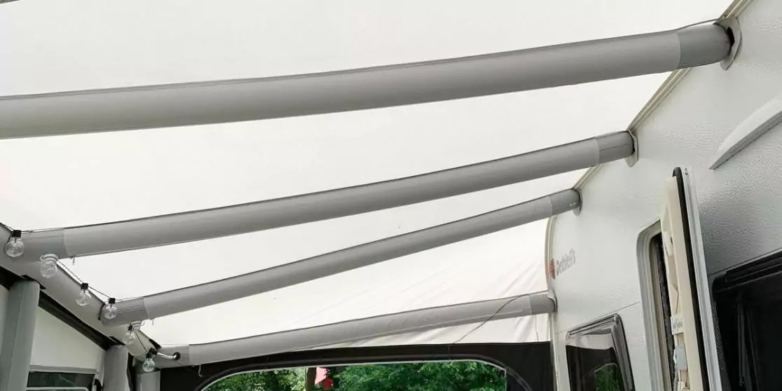 Blick unter wei&szlig;es Markisendach mit gebogenen Metallstreben, Seitenwand mit Fenster und T&uuml;r, gr&uuml;ne B&auml;ume im Hintergrund