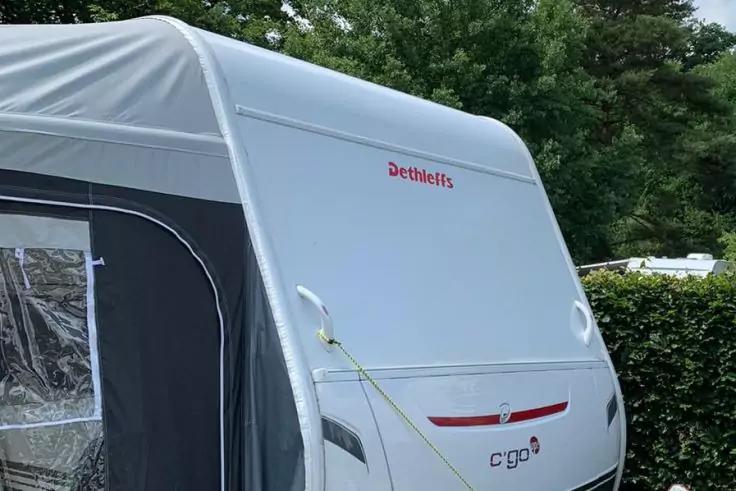 Vorderseite eines wei&szlig;en Wohnwagens mit grauem Vorzelt links, rotem Schriftzug "Dethleffs", Modellschriftzug "c'go", gelber Abspannleine, B&auml;ume und Hecke im Hintergrund, teilweise sichtbarer Nachbarwohnwagen rechts