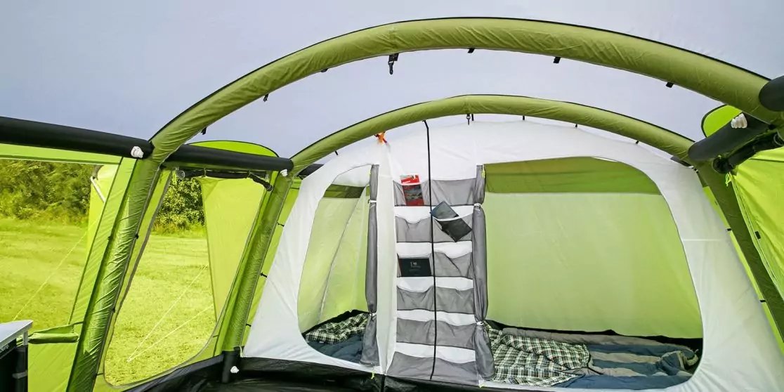 Innenraum eines gr&uuml;nen Zelts mit aufblasbaren Gest&auml;ngeb&ouml;gen, offenem zentralen Bereich, zwei getrennten Schlafbereichen mit Matratzen, h&auml;ngenden Aufbewahrungstaschen und Blick auf eine Wiese durch ein Zeltfenster
