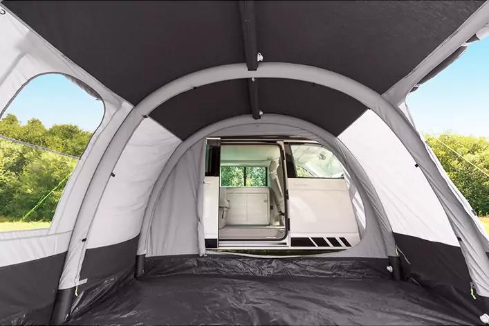 Blick aus dem Inneren eines großen Vorzelts auf einen weißen Camper mit geöffneter Schiebetür; Gras und Bäume außen