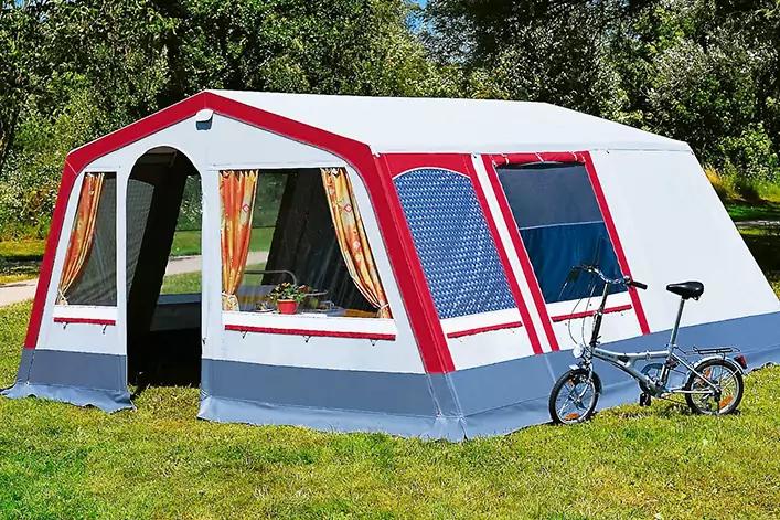 Großes weißes Zelt mit roten Rahmen, offener Eingang mit gelb-orangenen Vorhängen, zwei Fenster, Fahrrad lehnt am Zelt, Gras und Bäume im Hintergrund