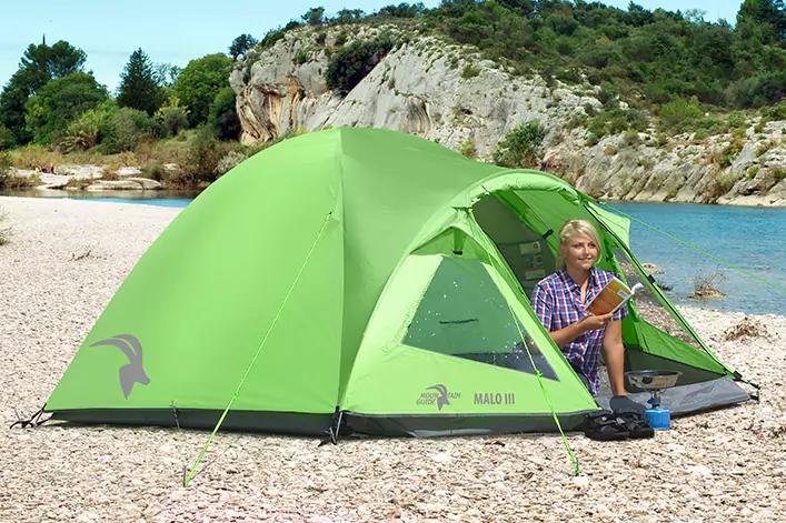 Grünes Campingzelt auf kiesigem Strand neben Wasser und Felsen, Person sitzt im geöffneten Eingang und hält ein Buch, Campinggeschirr neben der Person