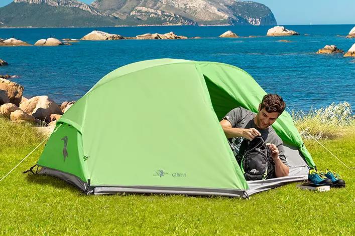 Zwei Personen in einem geöffneten grünen Zelt auf einer Wiese am Meer; Felsen im Wasser und Berge im Hintergrund; Rucksack und Schuhe neben dem Zelt