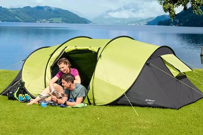 Zwei Personen sitzen vor einem grünen Tunnelzelt auf einer Decke am Ufer eines Sees; Berge und bewölkter Himmel im Hintergrund
