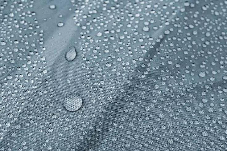 Wassertropfen unterschiedlicher Größe auf bläulich-grauem, strukturiertem Stoff mit sichtbarer Naht