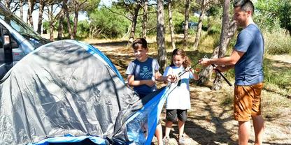 Ein Mann und zwei Kinder bauen ein blau-graues Zelt auf; im Hintergrund B&auml;ume und ein geparktes wei&szlig;es Fahrzeug