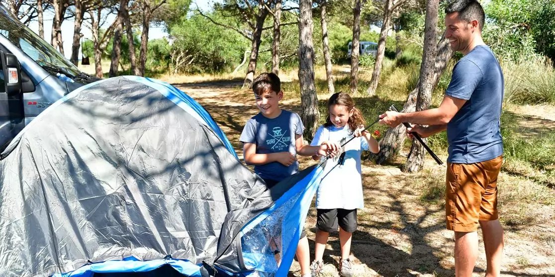 Ein Mann und zwei Kinder bauen ein blau-graues Zelt auf; im Hintergrund B&auml;ume und ein geparktes wei&szlig;es Fahrzeug