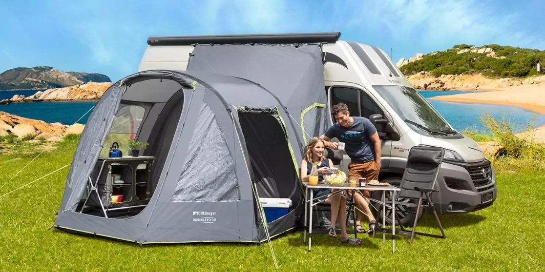 Wohnmobil mit angebautem Vorzelt auf gr&uuml;ner Wiese, zwei Personen an einem Campingtisch mit St&uuml;hlen, Geschirr und Bechern auf dem Tisch; im Hintergrund Meer, Sandstrand, Felsen und blauer Himmel