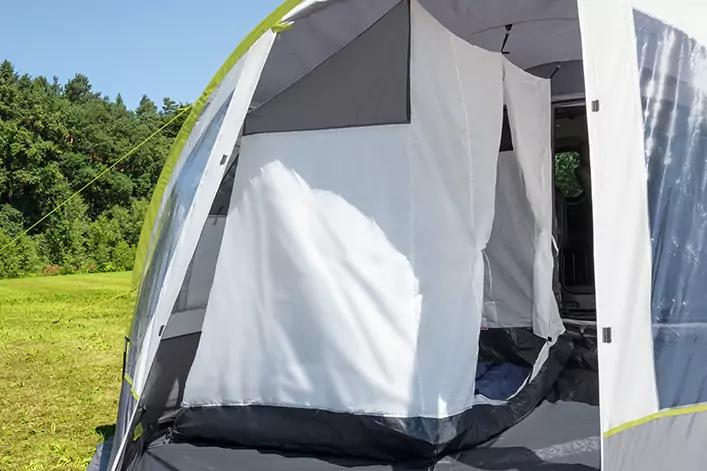 Zelt mit ge&ouml;ffneter Eingangst&uuml;r, wei&szlig;en h&auml;ngenden Innenw&auml;nden, grauem Boden, gr&uuml;nem Zeltkeder; Wiese und B&auml;ume im Hintergrund