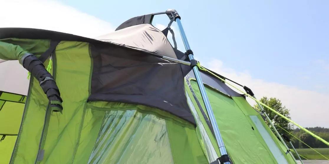 Close-up van een groene tent met een open raam en metalen stangen, blauwe lucht en bomen op de achtergrond