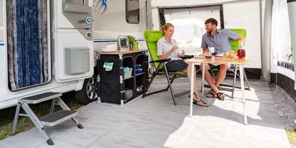 Zwei Personen sitzen an einem Klapptisch im Vorzelt eines Wohnwagens. Auf dem Tisch: Gl&auml;ser, eine Karaffe mit rotem Getr&auml;nk, Teller und Besteck. Campingst&uuml;hle, ein K&uuml;chenmodul mit Sp&uuml;le und die Einstiegsstufe des Wohnwagens sind sichtbar