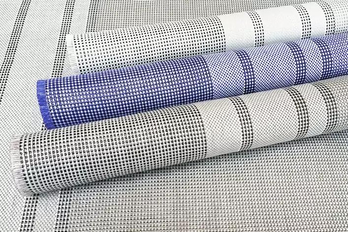 Drei aufgerollte Gewebe mit kariertem Muster (zwei helle mit schwarzem Raster, eines mit blauem Rand) liegen auf einer größeren gewebten Unterlage