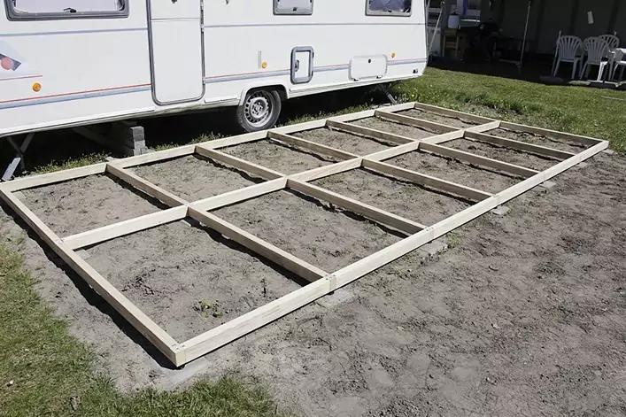 Holzrahmen mit Querverstrebungen auf nacktem Boden neben einem weißen Wohnwagen