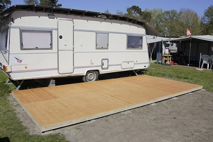 Weißer Wohnwagen auf einem Grasplatz, davor eine rechteckige Holzplattform; rechts ein ausgefahrener Vorbau mit Gartenstühlen und kleinem Tisch, Bäume im Hintergrund