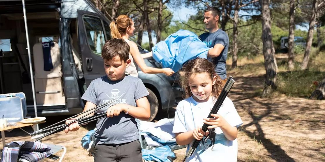 Zwei Kinder im Vordergrund halten Metallstangen; im Hintergrund heben zwei Erwachsene eine blaue Plane neben einem Wohnmobil in einem Kiefernwald