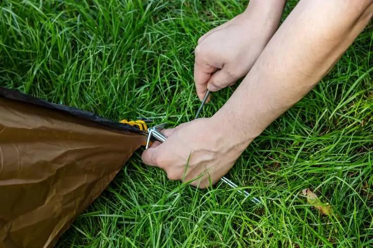 Zwei H&auml;nde dr&uuml;cken einen Hering durch eine &Ouml;se eines braunen Zelttuchs in gr&uuml;nes Gras