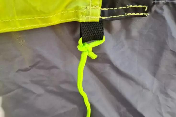 Nahaufnahme einer neongelben Kordel mit einfachem Knoten, befestigt an einem schwarzen Gurtst&uuml;ck unter einem neongelben Stoffstreifen auf grauem Stoff mit sichtbaren N&auml;hten