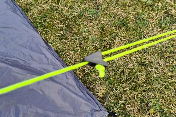 Ecke einer grauen Zeltplane mit neongelber Spannleine durch einen schwarzen Kunststoff-Spanner auf Gras