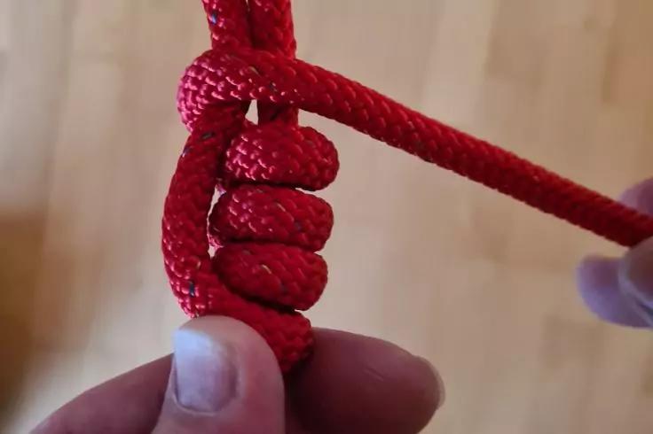 Nahaufnahme eines geflochtenen roten Seils mit mehreren eng gewickelten Schlaufen um einen festen Teil; zwei H&auml;nde halten das Seil; Holzfu&szlig;boden im Hintergrund