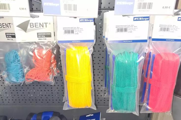 Mehrere transparente Plastikbeutel mit bunten Schn&uuml;ren h&auml;ngen an einem perforierten Metallblech; oben sind Preisschilder mit Barcodes sichtbar