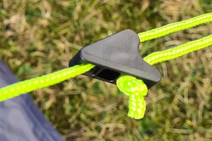 Zwarte driehoekige plastic klem op neongeel-groen koord met een kleine knoop; op de achtergrond gras en een stuk blauw stof