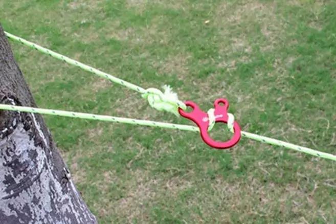 Baumstamm links, gr&uuml;ner Rasen im Hintergrund, zwei hellgr&uuml;ne Seile gespannt, roter Kunststoffclip an den Seilen, Knoten an einem Seil
